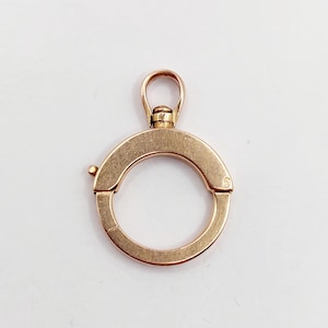 Può includere: Un fermaglio circolare color oro con un'apertura a cerniera e un piccolo anello nella parte superiore. Il fermaglio è progettato per aprirsi e chiudersi e ha un design semplice ed elegante. Lo sfondo è bianco.