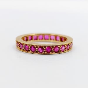 Könnte beinhalten: Ein goldener Eternity-Ring mit einer durchgehenden Reihe runder, leuchtend pinker Rubine, die in einer polierten Metallschiene gefasst sind. Der kreisförmige Ring zeigt eine gleichmäßige Anordnung der Edelsteine rundherum und eine glänzende Metalloberfläche.