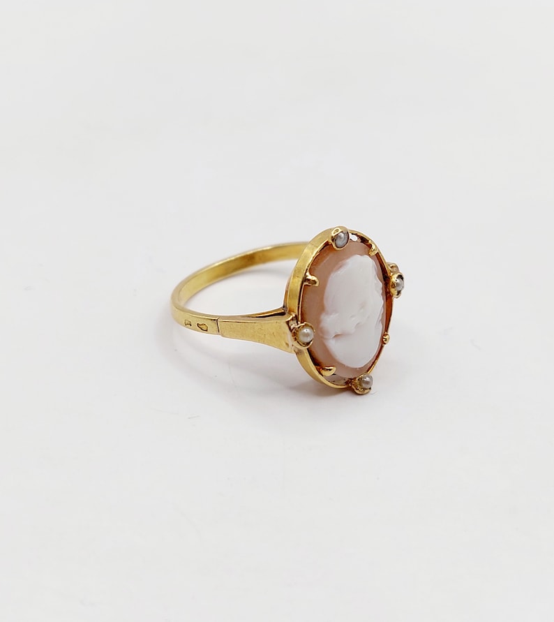May include: A gold ring with a cameo featuring a white profile portrait set in a shell-like background. The cameo is surrounded by small pearl-like stones.