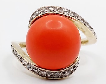 Può includere: Un anello d'oro con una grande gemma rotonda di colore arancione vibrante. La fascia si curva attorno alla pietra, impreziosita da piccoli diamanti scintillanti. L'anello è posto su uno sfondo bianco, evidenziando il suo design elegante.