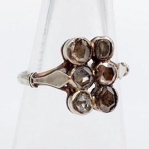 Op de afbeelding: Een zilveren ring met zeven lichtbruine, rond geslepen diamanten in een cluster. De ring heeft een gedetailleerde band en een decoratieve zetting. De ring wordt gepresenteerd op een helder, schuin oppervlak.