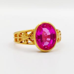 May include: A gold ring featuring a large, oval-cut, vibrant pink gemstone set in a bezel. The band displays an intricate, openwork filigree design with scrolling metal patterns that extend from the central setting along the sides of the ring.