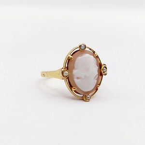 May include: A gold ring with a cameo featuring a woman's profile. The cameo is surrounded by a delicate gold frame and accented with small pearls.
