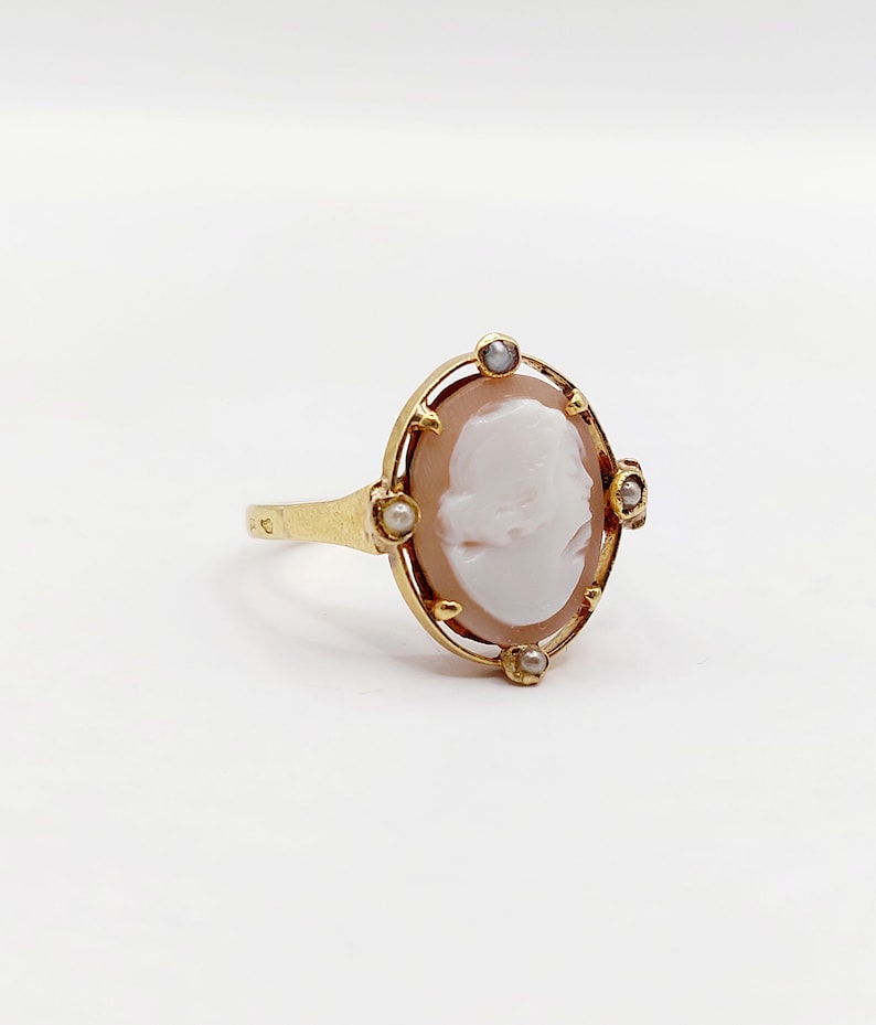 May include: A gold ring with a cameo featuring a woman's profile. The cameo is surrounded by a delicate gold frame and accented with small pearls.