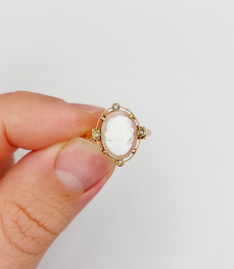 May include: A gold ring with a white cameo set in a gold frame with small pearl accents.