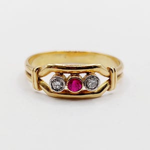 May include: A gold ring with a central red gemstone and two clear gemstones. The ring's design includes a distinctive band. The gemstones are set in a decorative setting. The ring is a piece of jewellery.