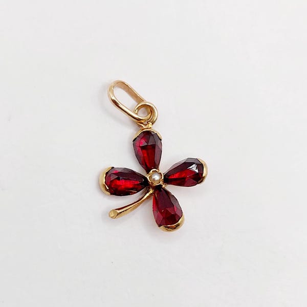 Pode incluir: Um pingente de ouro em formato de trevo de quatro folhas, apresentando quatro pedras preciosas granadas vermelho-escuras lapidadas em formato de pera, dispostas como pétalas ao redor de uma pequena pérola branca central, com uma argola dourada.