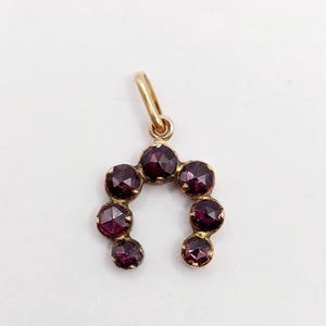 Op de afbeelding: Een hoefijzervormige hanger van goud met zeven ronde, gefacetteerde dieprode granaat edelstenen gezet in metaal. De hanger is voorzien van een klein gouden oogje aan de bovenzijde voor bevestiging aan een ketting, met een klassiek ontwerp.