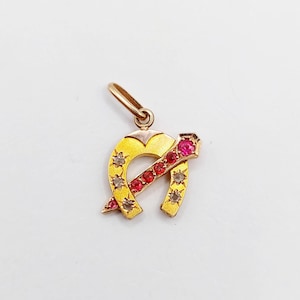 Peut inclure: Un pendentif en forme de fer à cheval en or avec une barre diagonale sertie de cinq pierres rondes rouges et quatre pierres rondes transparentes incrustées dans la surface dorée. Le pendentif comporte un petit anneau en or au sommet.