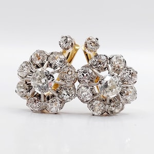 Op de afbeelding: Een paar bloemvormige oorbellen van geelgoud en zilver, elk voorzien van een centrale diamant in ronde slijpvorm omringd door een halo van acht kleinere diamanten, met een extra enkele diamant gezet op de steker boven het bloemmotief.