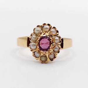 May include: A gold ring featuring a central oval-cut, deep pink gemstone surrounded by a halo of small, round pearls. The ring's band is a simple, polished gold. The setting has a floral design.