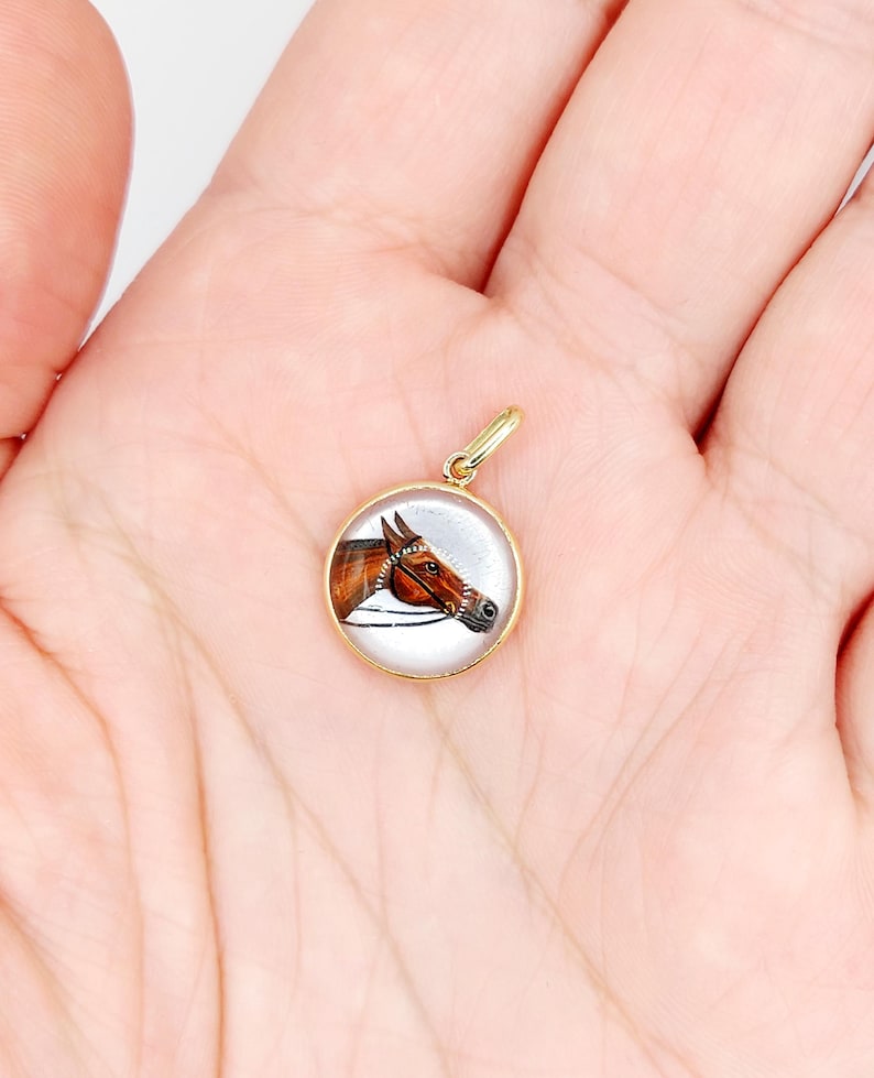 May include: A gold pendant with a round glass cabochon featuring a hand-painted portrait of a brown horse's head. The horse is wearing a bridle and has a white background.