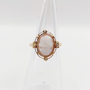 May include: A gold ring with a cameo featuring a woman's profile. The cameo is surrounded by a gold frame with small pearls.