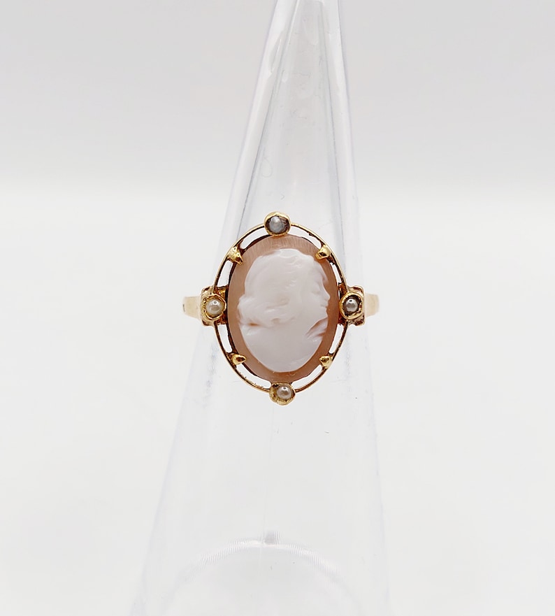 May include: A gold ring with a cameo featuring a woman's profile. The cameo is surrounded by a gold frame with small pearls.