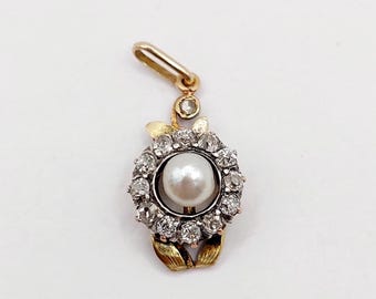May include: A gold pendant featuring a central white pearl surrounded by a circular halo of small clear stones. The pendant includes a small bail at the top with a single clear stone and two gold leaf-like accents at the bottom.