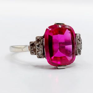 Puede incluir: Un anillo de tono plateado con una gran piedra preciosa de color rosa vibrante, de corte rectangular tipo cojín, engastada en el centro. La banda está decorada a ambos lados con pequeñas piedras claras de corte redondo en un engaste metálico.