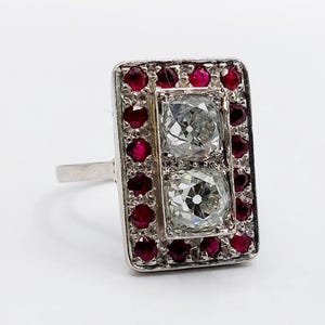 May include: A rectangular silver-toned ring featuring two central cushion-cut clear stones arranged vertically. The center stones are surrounded by a border of small, round, deep red stones set within the rectangular frame of the ring band.