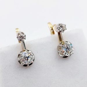 May include: A pair of earrings with a gold and silver setting. Each earring has a small diamond at the top and a larger, sparkling diamond at the bottom. The earrings are displayed against a white background.
