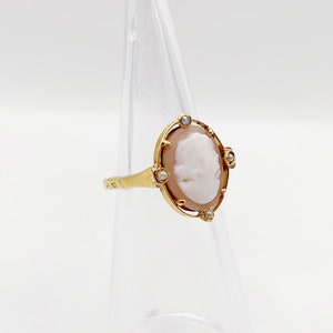 May include: A gold ring with a cameo featuring a woman's profile. The cameo is surrounded by a gold frame with four small pearl accents.