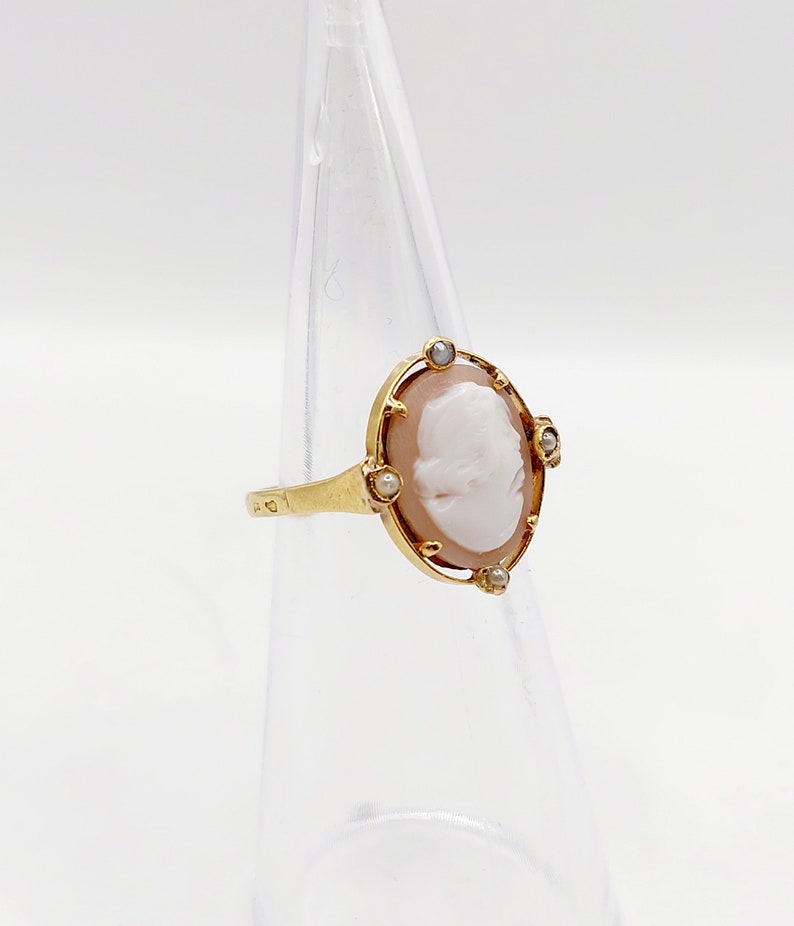 May include: A gold ring with a cameo featuring a woman's profile. The cameo is surrounded by a gold frame with four small pearl accents.