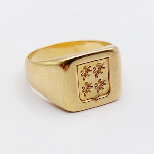May include: A gold signet ring featuring a square face with an engraved shield design. The shield contains four small, identical lion figures arranged in a two-by-two grid pattern. The ring has a polished, smooth finish and a solid band construction.