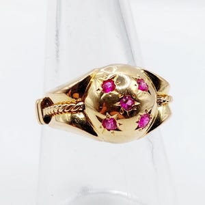 May include: A gold ring with a rounded center, featuring five small, bright red gemstones set within star-shaped designs. The ring has a textured band and a twisted rope detail on the sides, showcasing a vintage aesthetic.