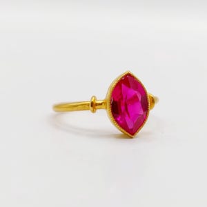 May include: A gold ring featuring a marquise-cut vibrant pink gemstone set in a beaded metal bezel. The band is smooth and round, connecting to the central stone setting with small decorative metal accents on each side of the gemstone.