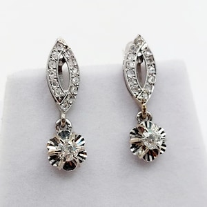 May include: A pair of silver-toned drop earrings featuring a marquise-shaped top section encrusted with small clear stones and a dangling flower-shaped charm at the base, which is set with a single larger clear stone in the centre of the petals.