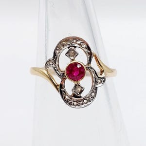 May include: A gold ring featuring a central round red gemstone set in a yellow gold bezel. The centerpiece is surrounded by an oval-shaped frame made of silver-toned metal, accented with small embedded stones and two diamond-shaped details.