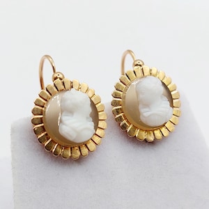 Op de afbeelding: Een paar gouden oorbellen met een rond, beige-kleurig camee-middelpunt met een wit, uitgesneden profiel. Het camee is omringd door een geschulpte, goudkleurige rand in de vorm van zonnestralen en bevestigd aan een gebogen gouden haakje.