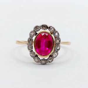 Könnte beinhalten: Ein goldener Ring mit einem ovalen, leuchtend roten Rubin im Zentrum, umgeben von einem Kranz aus kleinen, funkelnden Diamanten. Der Ring hat ein schlichtes Band, das die Edelsteine zur Geltung bringt.