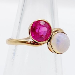 Könnte beinhalten: Ein goldener Ring mit einem großen, runden, facettierten Rubin und einem kleineren, runden, irisierenden Mondstein. Die Edelsteine sind in Goldfassungen gefasst, und der Ring hat einen zarten, gebogenen Ring.