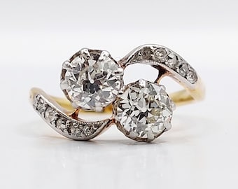 Può includere: Un anello bypass con due pietre rotonde prominenti incastonate in metallo color argento, accentuato da pietre più piccole lungo la fascia curva. L'anello è realizzato con un gambo color oro che sostiene l'incastonatura a due pietre.