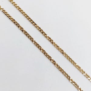 May include: Two gold-colored chain necklaces are diagonally positioned against a white background. The chains have a curb link design, with each link appearing to be flat and slightly rounded. The chains are close together, creating a parallel effect.
