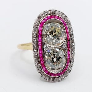 May include: An oval-shaped ring featuring two large, clear diamonds. The diamonds are surrounded by a row of small, pink gemstones and a border of smaller, clear stones. The band is gold-colored.