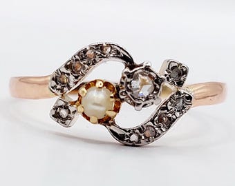 May include: A delicate antique ring with a pearl and two diamonds set in a gold and silver band. The ring features a unique, swirling design.