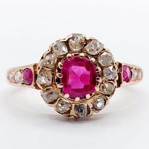 Puede incluir: Un anillo vintage de oro rosa con un gran rubí rodeado por un halo de diamantes más pequeños. El anillo tiene una banda delicada con rubíes y diamantes más pequeños.