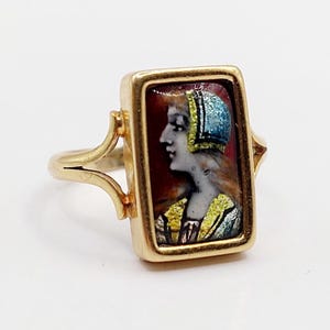 Puede incluir: Anillo de oro con una cara rectangular que muestra un retrato de una mujer de perfil. El retrato tiene un fondo rojo, y la mujer lleva un tocado y ropa azules y amarillos. El anillo tiene un diseño delicado y ornamentado.