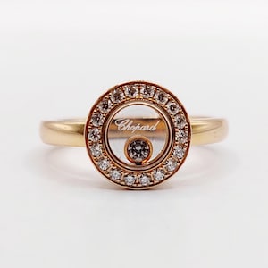 Peut inclure: Une bague en or avec un design circulaire. Le centre présente un espace clair avec le nom de la marque "Chopard" et un diamant flottant. Le cercle extérieur est incrusté de petits diamants. La bague est posée sur un fond blanc.