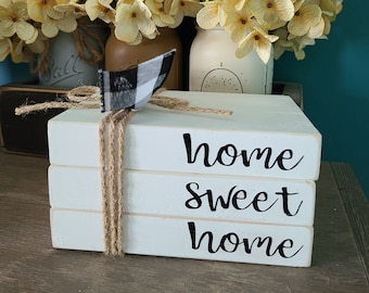 Op de afbeelding: Een stapel van drie witte, beschilderde houten blokken met de woorden "home sweet home" in het zwart. De blokken zijn aan elkaar gebonden met touw en een zwart-wit geruit lint. De blokken staan op een houten oppervlak voor drie glazen potten gevuld met gele bloemen.