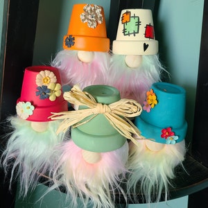 Op de afbeelding: Een verzameling handgemaakte kabouterbeeldjes. Elke kabouter heeft een beschilderde terracotta pottenhoed in verschillende kleuren, waaronder oranje, rood en blauwgroen, versierd met bloemmotieven. De kabouters hebben pluizige, pastelkleurige baarden en houten neuzen.