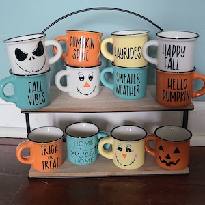 May include: A set of 10 ceramic mugs with various fall-themed designs. The mugs are arranged on a two-tiered metal stand. The designs include "Pumpkin Spice", "Hayrides", "Happy Fall", "Sweater Weather", "Hello Pumpkin", "Fall Vibes", "Trick or Treat", "Home Sweet Home", and two mugs with cute faces.
