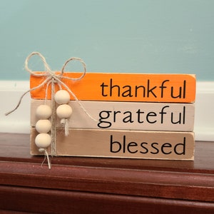 Puede incluir: Una pila de tres bloques de madera pintados en naranja, beige y marrón claro. Los bloques están apilados uno encima del otro y tienen las palabras "thankful", "grateful" y "blessed" escritas en negro. Una cadena de cuentas de madera cuelga del bloque superior.