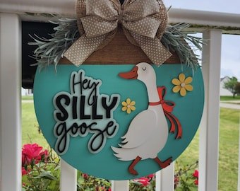 May include: A round, teal wooden sign with the words "Hey Silly Goose" and a cartoon goose. The sign is decorated with a burlap bow, greenery, and yellow flower accents. It is hanging on a white railing with red roses in the foreground.