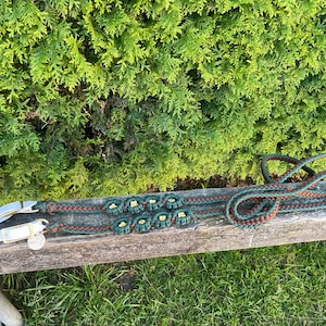 May include: A green and brown braided leather horse rein with gold metal accents. The rein is lying on a wooden surface.