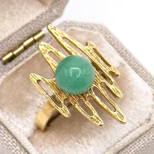 May include: A gold-toned ring featuring a central round green stone set against an abstract, textured metal frame with elongated, jagged prongs that extend outward from the center, creating a distinct layered appearance on the band.