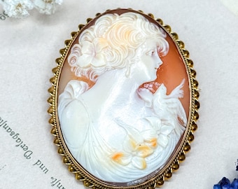 May include: Oval cameo brooch featuring a woman's profile carved in white against an orange background. The brooch has a gold-colored, ornate frame. The woman is wearing a dress and holding a dove. The brooch is a vintage jewelry piece.