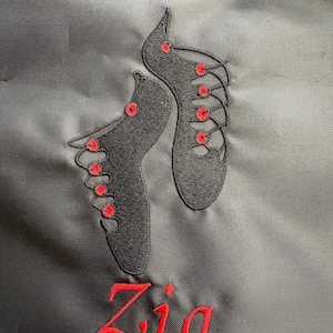 May include: A black fabric surface featuring an embroidered design of two black ghillie shoes with red eyelets and laces. Below the shoes, the name Zia is embroidered in red script lettering. The embroidery is detailed with black and red thread.