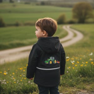 May include: A child in a black hooded jacket with a green and yellow tractor motif on the back, standing in a field. The jacket features a reflective stripe. The child wears dark blue jeans and grey trainers.