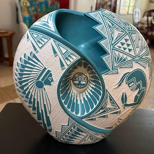 May include: A round, teal and white ceramic vase featuring intricate etched designs. The surface displays a profile of a figure in a feathered headdress, geometric patterns, and a central circular medallion with a stylized face motif.